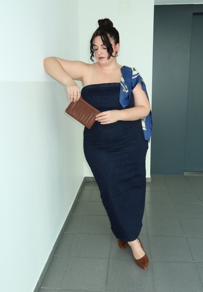 Vestido maxi de mezclilla en azul oscuro con silueta ajustada, combinado con un clutch marrón y una bufanda azul con patrón. Tacones marrones de punta.