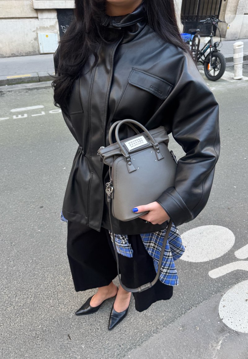 Personne portant une veste en cuir noire et des chaussures pointues noires, tenant un sac à main gris avec un détail en tissu bleu à carreaux, debout dans la rue.