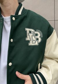 Vihreä villasekoite varsity-takki, jossa on kermaiset nahkaiset hihat, suuri kirjailtu logo ja raidalliset resorit hihansuissa.