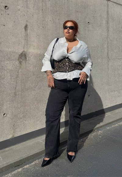 Persona con blusa blanca con corsé negro, vaqueros negros y zapatos negros puntiagudos, de pie contra una pared de hormigón a la luz del sol.