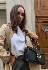 Sac à main en cuir noir avec quincaillerie dorée, forme structurée, surface texturée et anse. Modèle portant un manteau beige et un haut blanc devant un fond en bois.