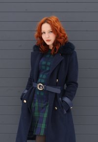 Femme aux cheveux rouges bouclés, portant un manteau bleu marine avec col en fourrure, une robe à carreaux verts et bleus, debout devant un mur gris à panneaux horizontaux.