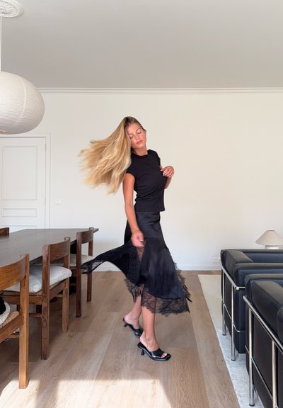 Top negro ajustado, falda negra fluida con borde de encaje y tacones abiertos negros. Mesa de comedor de madera y sofá de cuero en una habitación luminosa.