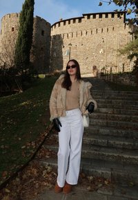 Abrigo de piel sintética color crema, suéter marrón claro, pantalones blancos de pierna ancha, guantes negros y botas marrones. De pie sobre escalones de piedra con un castillo de fondo.