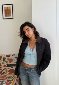 Una giovane donna con capelli scuri indossa un crop top blu chiaro, una giacca scura e jeans, in piedi accanto a un divano con motivo floreale e opere d'arte incorniciate.