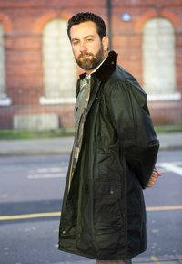 Dark green waterproof jacket featuring a corduroy collar, a silver zip, and two front pockets, worn over a patterned shirt and trousers.