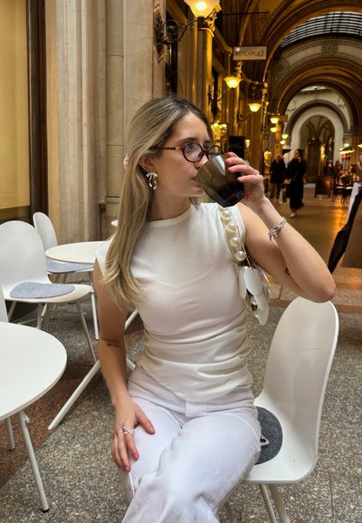 Top blanco con patrón texturizado y grandes perlas falsas en el hombro, combinado con pantalones blancos. Sosteniendo una taza de vidrio negro en un ambiente de café.