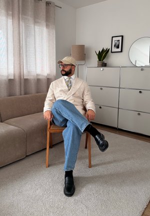 Hombre con blazer beige, gorra, corbata a rayas, gafas y vaqueros sentado con las piernas cruzadas en una silla de madera en una sala de estar moderna.