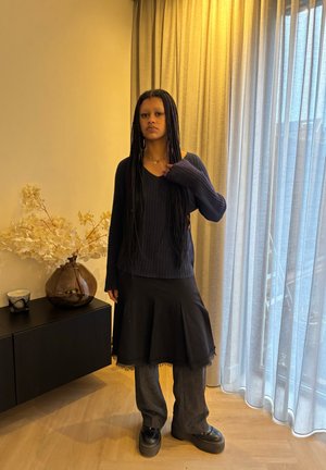 Suéter negro de canalé con escote en V, superpuesto sobre una falda negra con dobladillo deshilachado, combinado con pantalones grises anchos y zapatos negros gruesos.