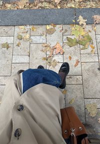 Béžový trenčkot, modré džínsy, čierne loafery a hnedá kabelka na kamennej dlažbe so zmiešanými jesennými listami.