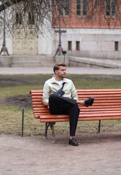 Hombre sentado en un banco de madera, vistiendo una chaqueta con cremallera gris clara, pantalones negros y zapatos negros, con un fondo arquitectónico desenfocado.