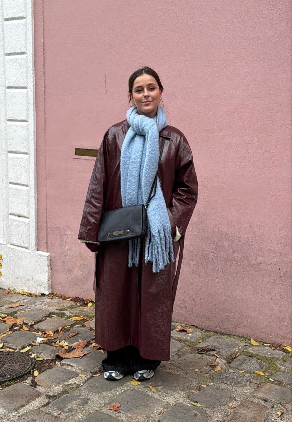 Jeune femme portant un long manteau bordeaux, une écharpe bleu clair, un sac bandoulière noir et des baskets, debout sur une rue pavée avec un mur rose en arrière-plan.