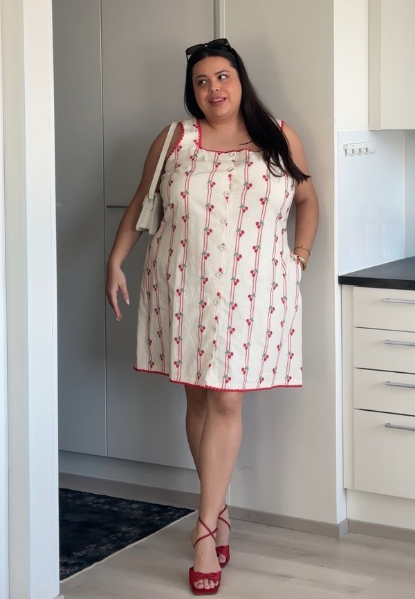 Femeie în rochie albă fără mâneci cu model de cireșe roșii, purtând o geantă albă de umăr, stând în ușa unei bucătării moderne.