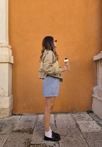 Mujer con pantalones cortos a rayas y chaqueta beige sosteniendo un cono de helado, de pie contra una pared texturizada naranja sobre un pavimento de piedra.