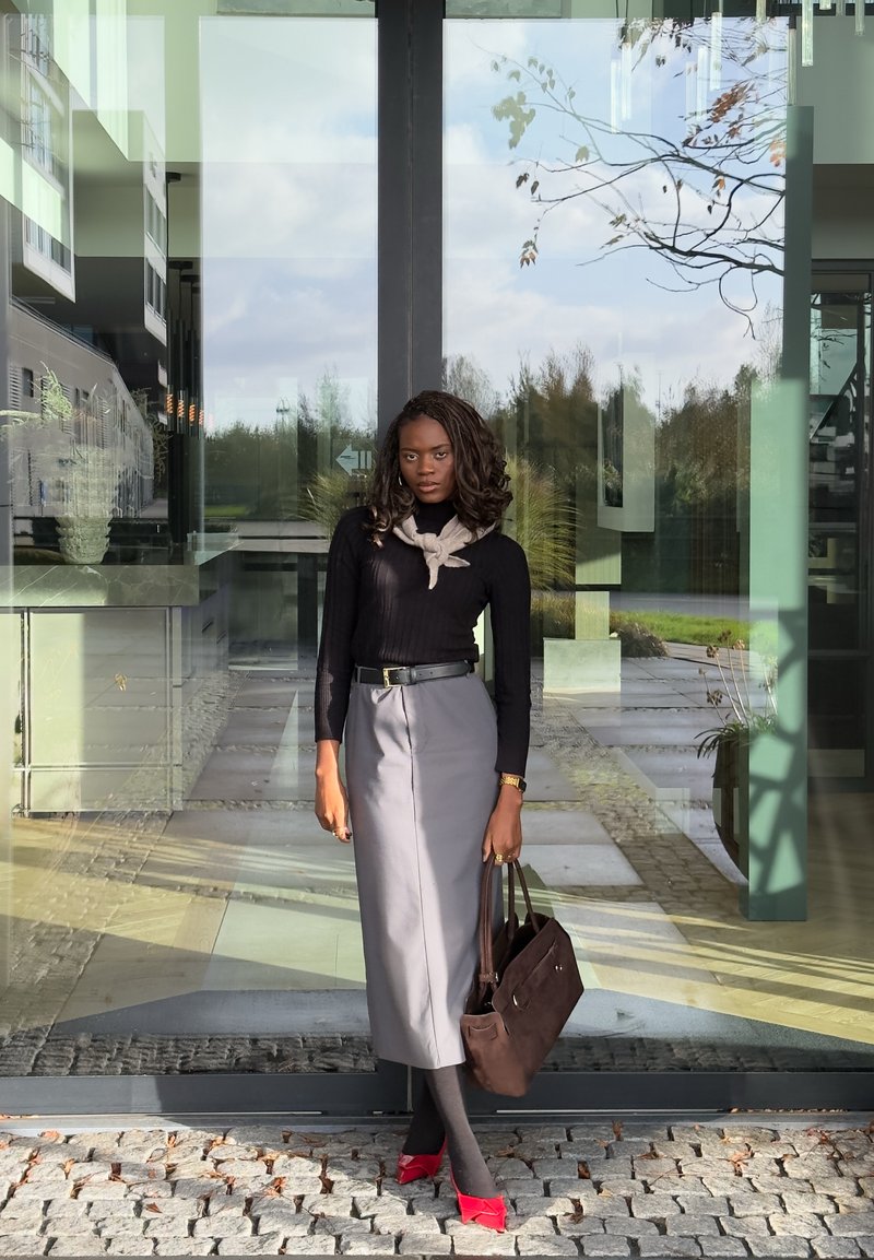 Mujer con blusa negra, falda midi gris y tacones rojos se encuentra sobre adoquines frente a puertas de cristal sosteniendo un bolso marrón.