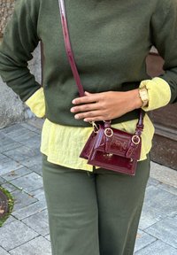 Burgundy patent leather handbag featuring two compartments, gold hardware accents, and a long adjustable strap, styled with a green sweater and a yellow blouse.