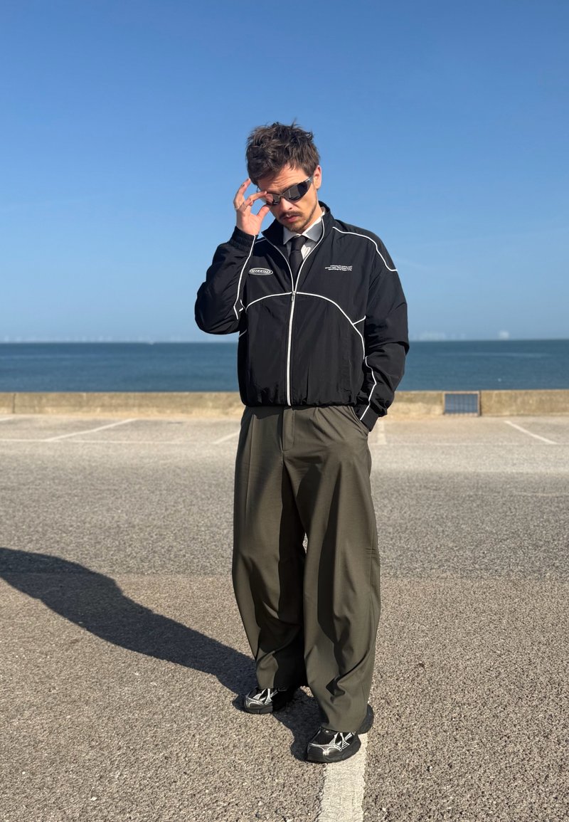 Mann med mørke solbriller, svart jakke, løse olivengrønne bukser og joggesko står på et steinlagt område nær sjøen under klar himmel.