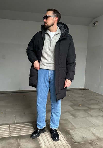 Abrigo de plumas negro, suéter gris, jeans azul claro y mocasines negros. El abrigo de plumas cuenta con capucha y diseño acolchado, usado en un entorno al aire libre.