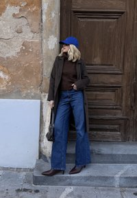 Pull marron, long manteau gris, jean denim à jambes larges, talons pointus marron et casquette bleue ; sac à main noir avec bandoulière. Fond de mur texturé.
