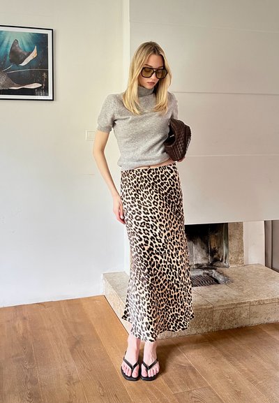 Top corto de punto gris de manga corta, falda deslizante de estampado de leopardo, chanclas negras, sosteniendo un bolso de mano acolchado marrón. El fondo presenta una chimenea.