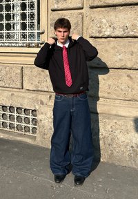 Fekete kapucnis felső, fehér inggel, piros csíkozott nyakkendővel és túlságosan nagy kék farmernadrággal. Fekete cipők egészítik ki az öltözetet. Városi háttér.