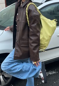 Veste en cuir marron coupe oversize, jeans baggy bleu clair et sac jaune à cordon de serrage. Chaussures blanches et marron à motifs.