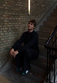 Svart langermet topp og vide bukser, sittende på steintrapp. Mørk vegg med en dekorativ lampe. Grønn og hvit slip-on sko.