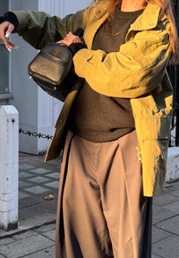 Olivgrüne Jacke, schwarzer Pullover und lockere braune Hose. Schwarze, strukturierte Handtasche in der rechten Hand, Zigaretten in der linken.