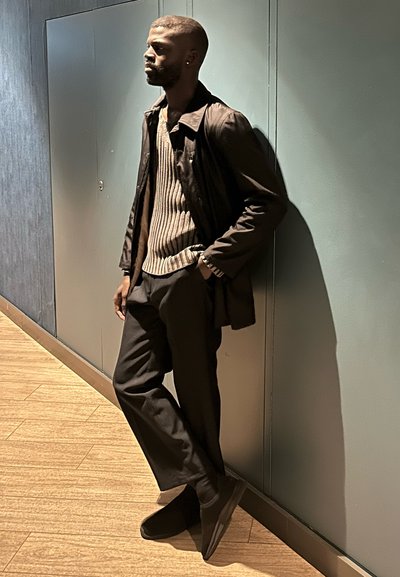 Hombre con cabello corto y barba apoyado contra una pared gris, vestido con una chaqueta oscura, suéter de rayas, pantalones negros y zapatos negros en el interior.