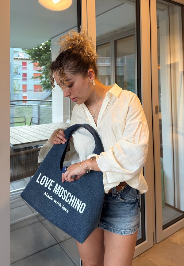 Ung kvinna med lockigt hår i vit blus och jeansshorts håller en blå Love Moschino-väska nära glasdörrar.