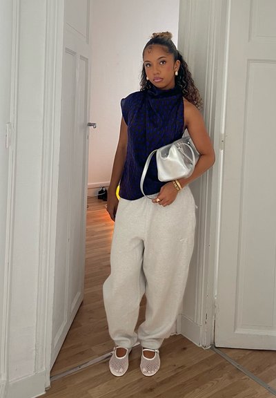 Top sin mangas de color azul marino con estampado, pantalones deportivos holgados blancos, zapatos de malla blancos tipo slip-on, sosteniendo un bolso brillante de color plata. Suelo de madera y marco de puerta visibles.