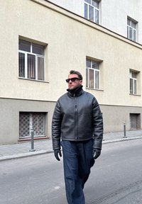 Άνδρας που φοράει μαύρο δερμάτινο μπουφάν, γάντια και γυαλιά ηλίου περπατώντας σε δρόμο με μπεζ κτίριο και παράθυρα στο φόντο.