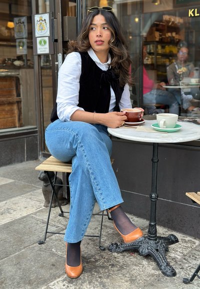 Persona vestida con un chaleco acolchado negro sobre una camisa blanca, jeans azules y tacones marrones, sentada en una mesa de mármol con tazas de café.