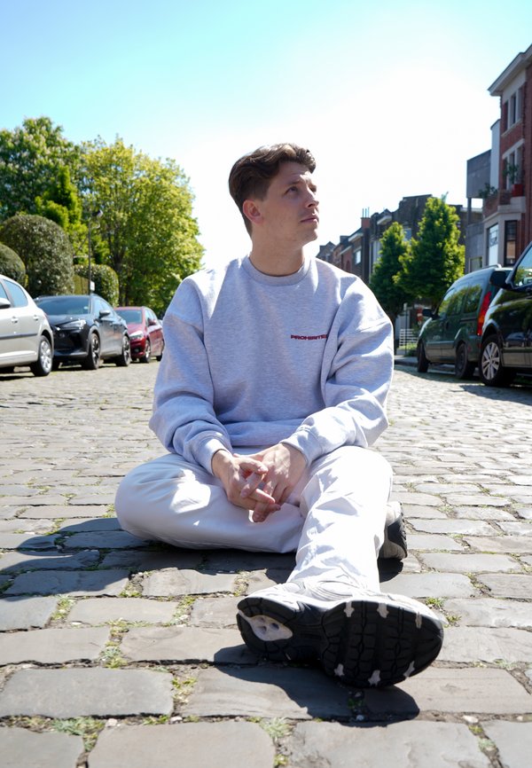 Sudadera de cuello redondo gris claro con texto rojo, combinada con pantalones blancos. Sentado con las piernas cruzadas en una calle de adoquines, con zapatillas negras visibles.