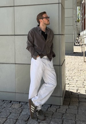 Hombre con gafas de sol, camisa oscura, pantalones blancos y zapatillas apoyado contra una pared de azulejos en un camino empedrado cerca de una bicicleta aparcada.