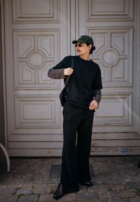 Homme en pantalon noir à jambes larges, portant un t-shirt à manches longues superposé à un t-shirt à manches courtes, casquette, lunettes de soleil et sac d'épaule, debout devant une porte ornée.