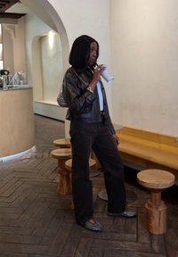 Braune Lederjacke über einem weißen Oberteil, dunklen Hosen und schwarzen Schuhen mit silbernen Nieten. Hält eine weiße Tasse. Holz- und glatte Oberflächen im Hintergrund.