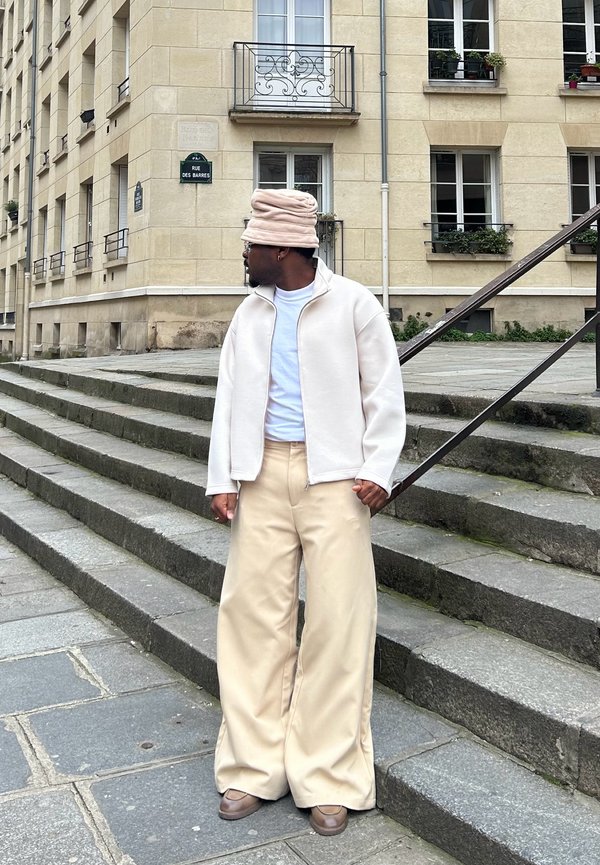 Veste beige surdimensionnée zippée, t-shirt blanc, large pantalon beige, et un chapeau marron clair, debout sur des marches en pierre près d'un bâtiment.