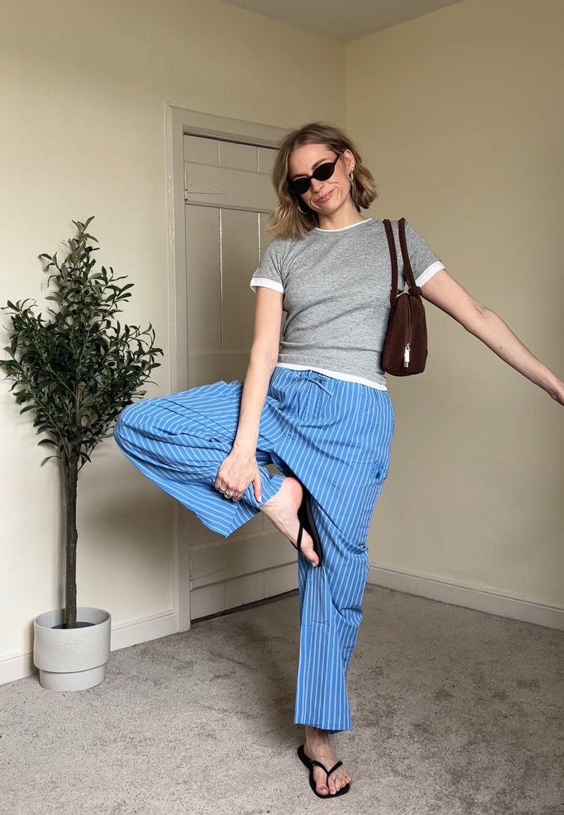 Frau mit Sonnenbrille, grauem T-Shirt, blau gestreifter Hose, hält eine braune Tasche und balanciert auf einem Bein in einem beigen Raum mit einer hohen Topfpflanze.