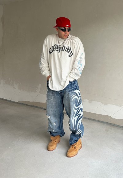Camisa blanca de manga larga con texto gráfico negro, jeans azules desgastados con un patrón tribal blanco y botas marrones. Gorra roja y gafas de sol.
