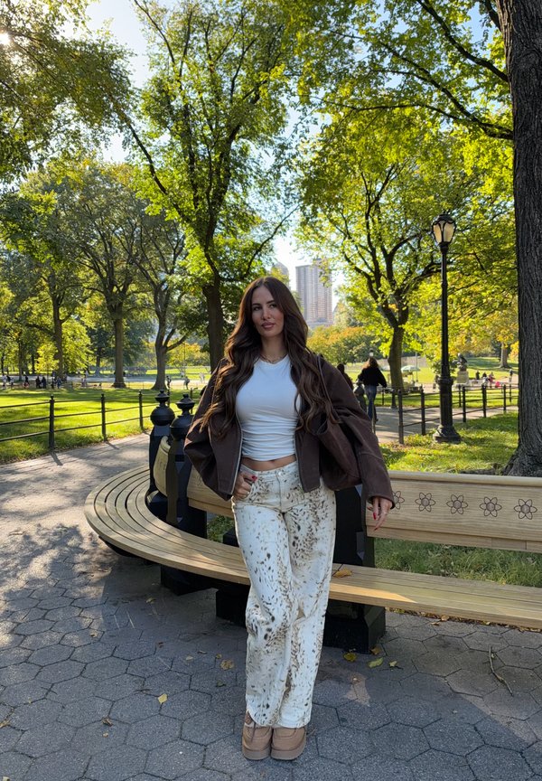 Una mujer está de pie junto a un banco curvo de madera en un frondoso parque urbano, con una farola y edificios lejanos detrás de ella.