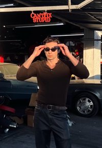 Férfi alak fekete hosszú ujjú ingben és fekete bőr nadrágban, napszemüveget igazítva, egy garázsban, ahol vintage autók és egy felirat található.