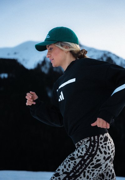 Sudadera negra con rayas blancas, gorra verde y leggings estampados en beige con manchas negras. Modelo corriendo en un paisaje nevado.