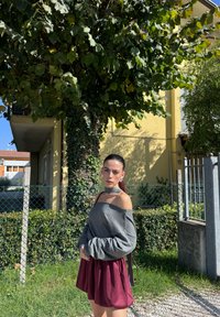 Grå tröja med off-shoulder-design och långa ärmar, tillsammans med en burgunderfärgad skaterkjol. Bakgrunden visar grönska och en gul byggnad.