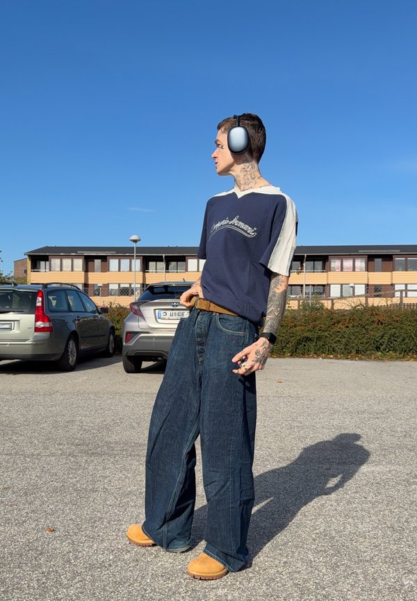 Un hombre tatuado con auriculares, una camisa azul marino, vaqueros de pernera ancha y zapatos de color tostado está de pie en un aparcamiento soleado.