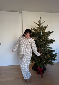 Beige kockás pizsamaszett kis fekete fenyőfa mintákkal, gombos felsővel és hozzá illő nadrággal. Kényelmes, texturált anyag. Közeli karácsonyfa.