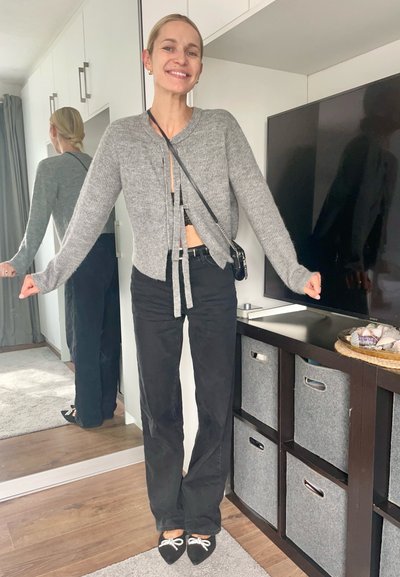 Cardigan gris corto con lazo en la parte delantera, combinado con pantalones negros de cintura alta y bailarinas negras con un lazo blanco. Elementos de fondo neutro.