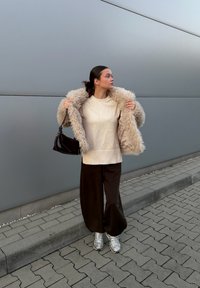 Beige faux fur coat worn over a cream jumper, teamed with loose brown trousers and silver trainers. Holding a black handbag, standing against a grey background.