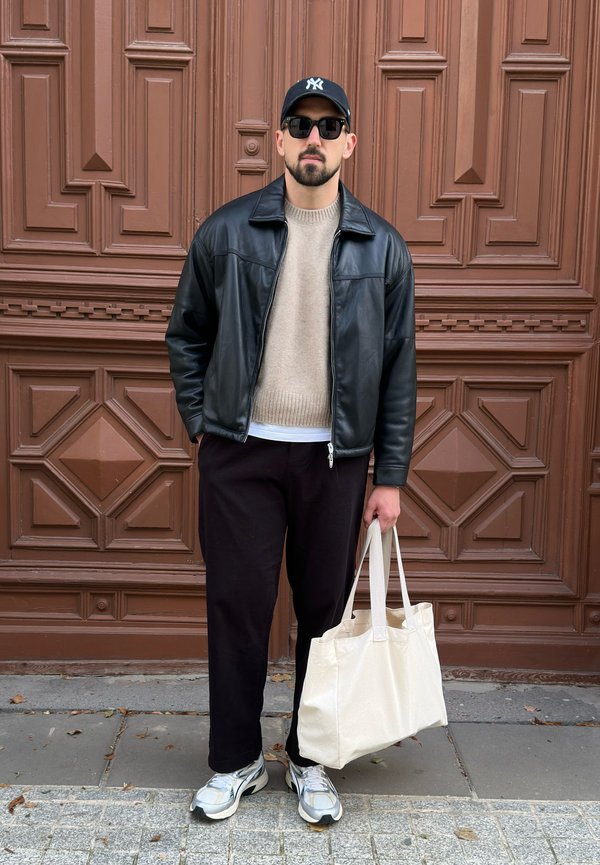 Giacca di pelle nera sopra un maglione beige, pantaloni scuri e sneakers grigie. Una borsa tote beige in mano, in piedi davanti a uno sfondo texture marrone.