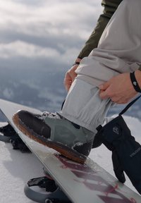 Snowboardstøvel med svart og grønn overdel, gummisåle og snøringer. Snødekket overflate og grå bukser med elastiske mansjetter i bildet.